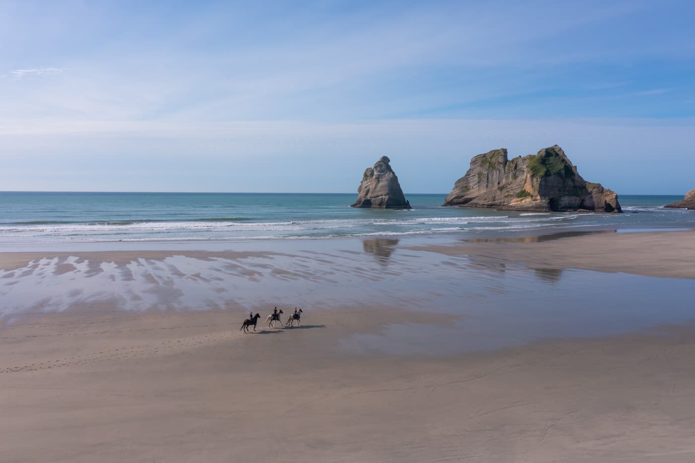 People horse riding along a wide sandy beach with sea and hills in Golden Bay, New Zealand